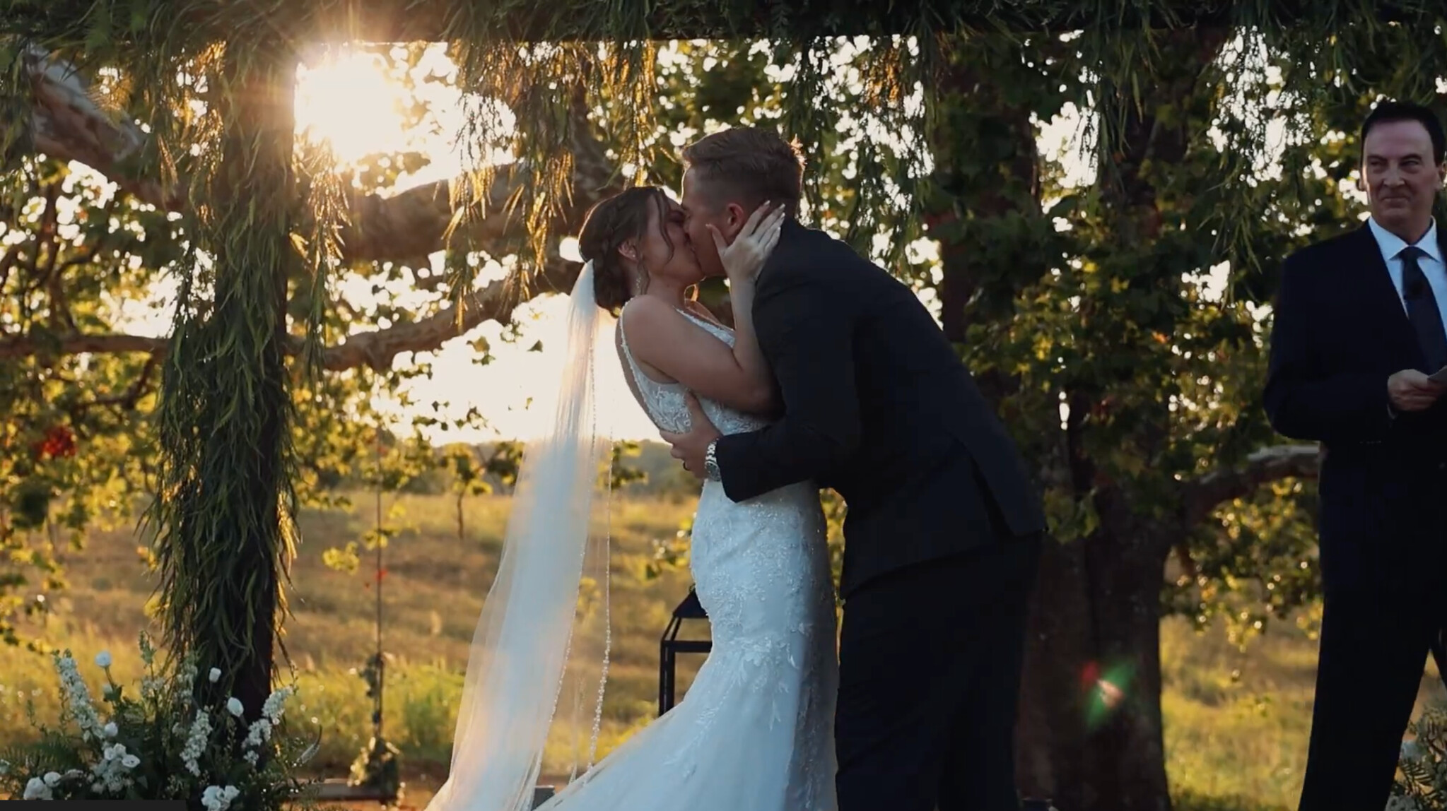 Aleah and Sam sharing their first kiss beneath a sunlit outdoor wedding arbor